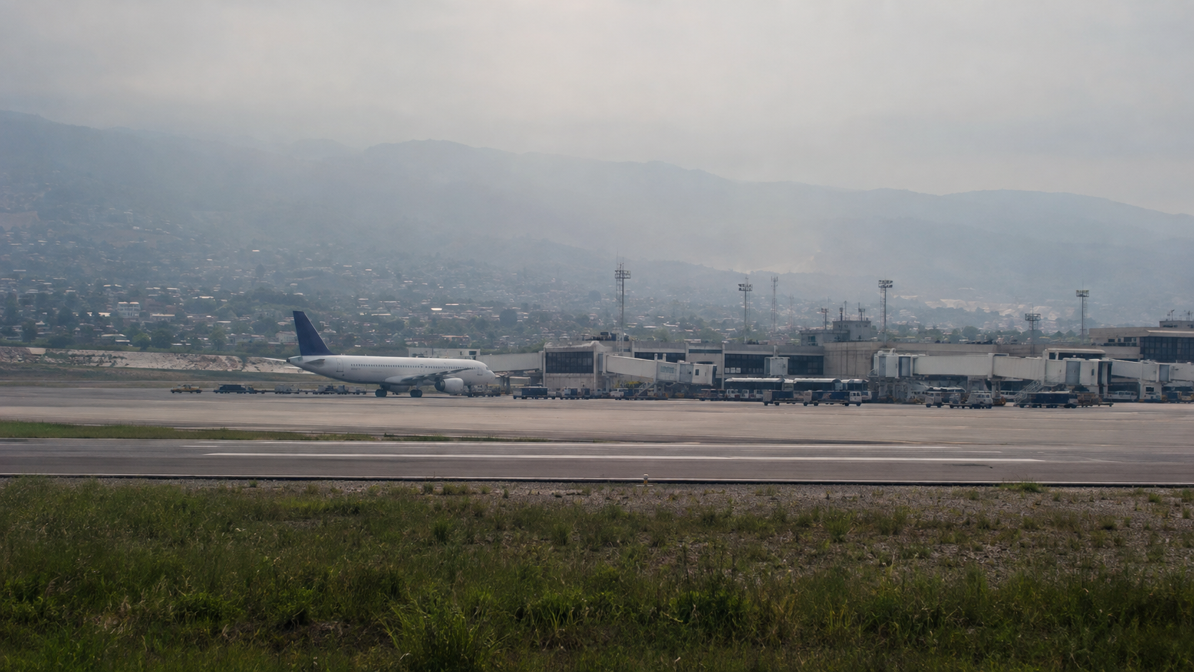 Editorial illustration of a grounded passenger aircraft at a tropical airport beneath hazy mountains