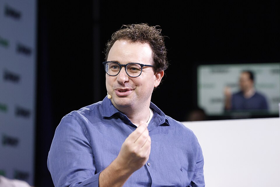 Dario Amodei speaks onstage in glasses and a blue shirt, wearing a headset microphone at TechCrunch Disrupt in 2023.