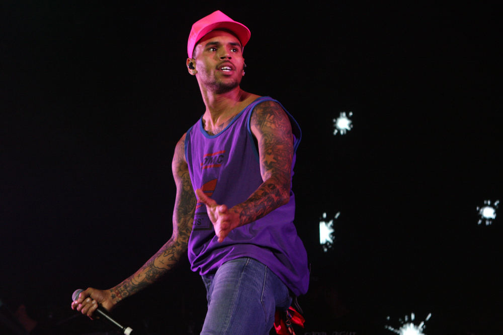 Chris Brown performing in a purple sleeveless shirt and bright pink cap, holding a microphone on a dark stage