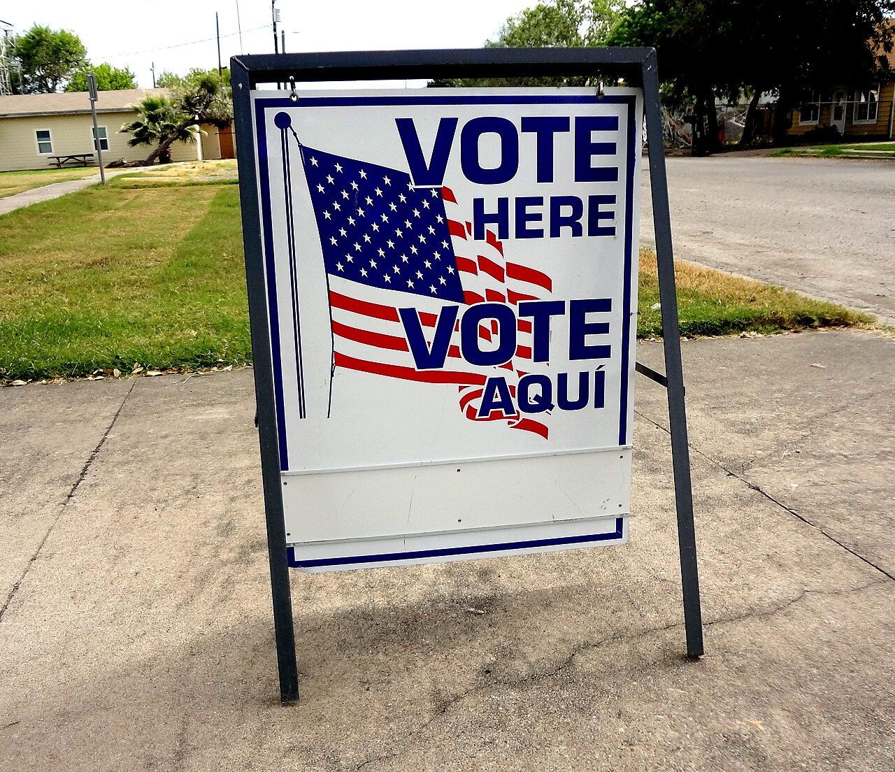 A freestanding red, white and blue sign reading Vote Here and Vote Aqui beside a street in Taft, Texas