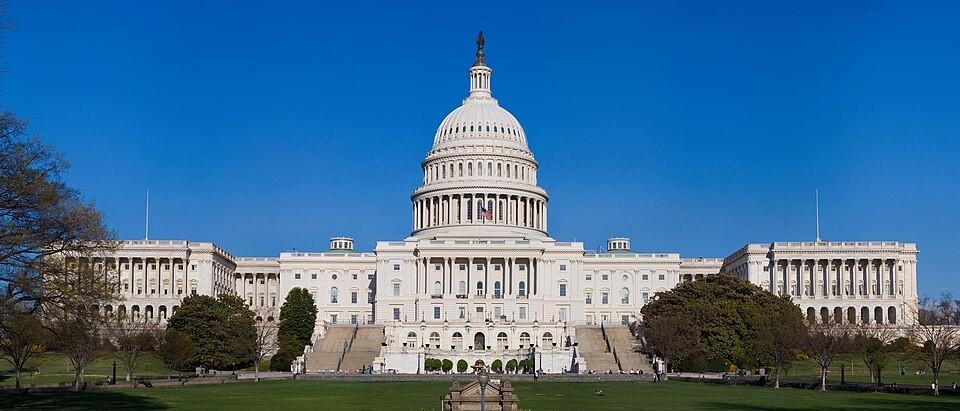 The western front of the U.S. Capitol, with its central dome, green lawn and trees beneath a clear blue sky