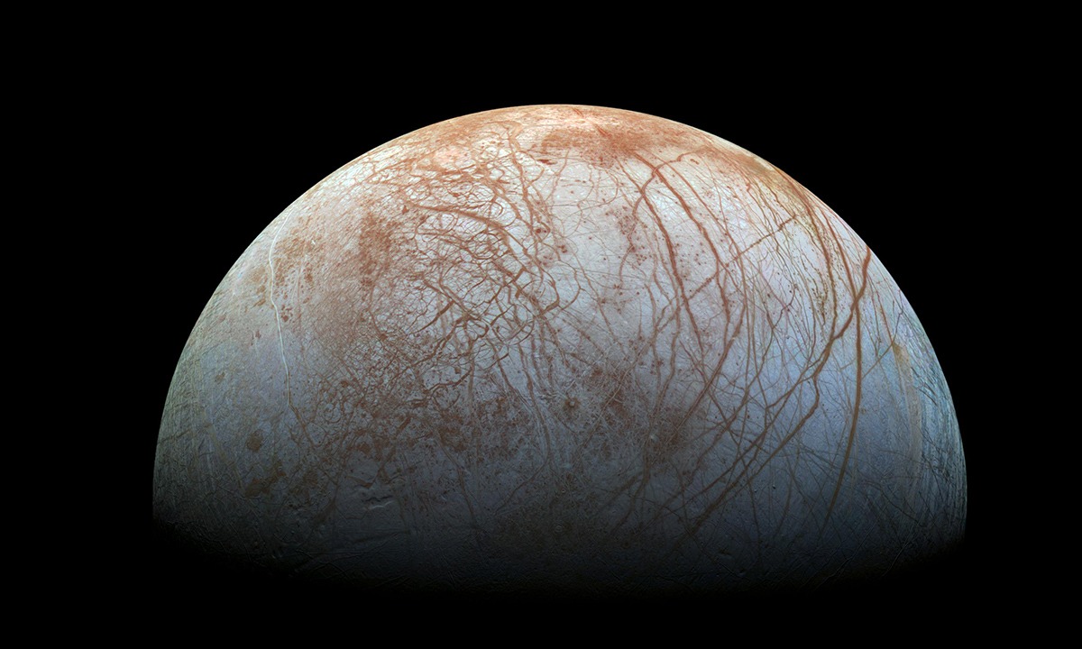 Processed-color Galileo photographic mosaic showing Europa’s pale icy surface crossed by reddish-brown lines.