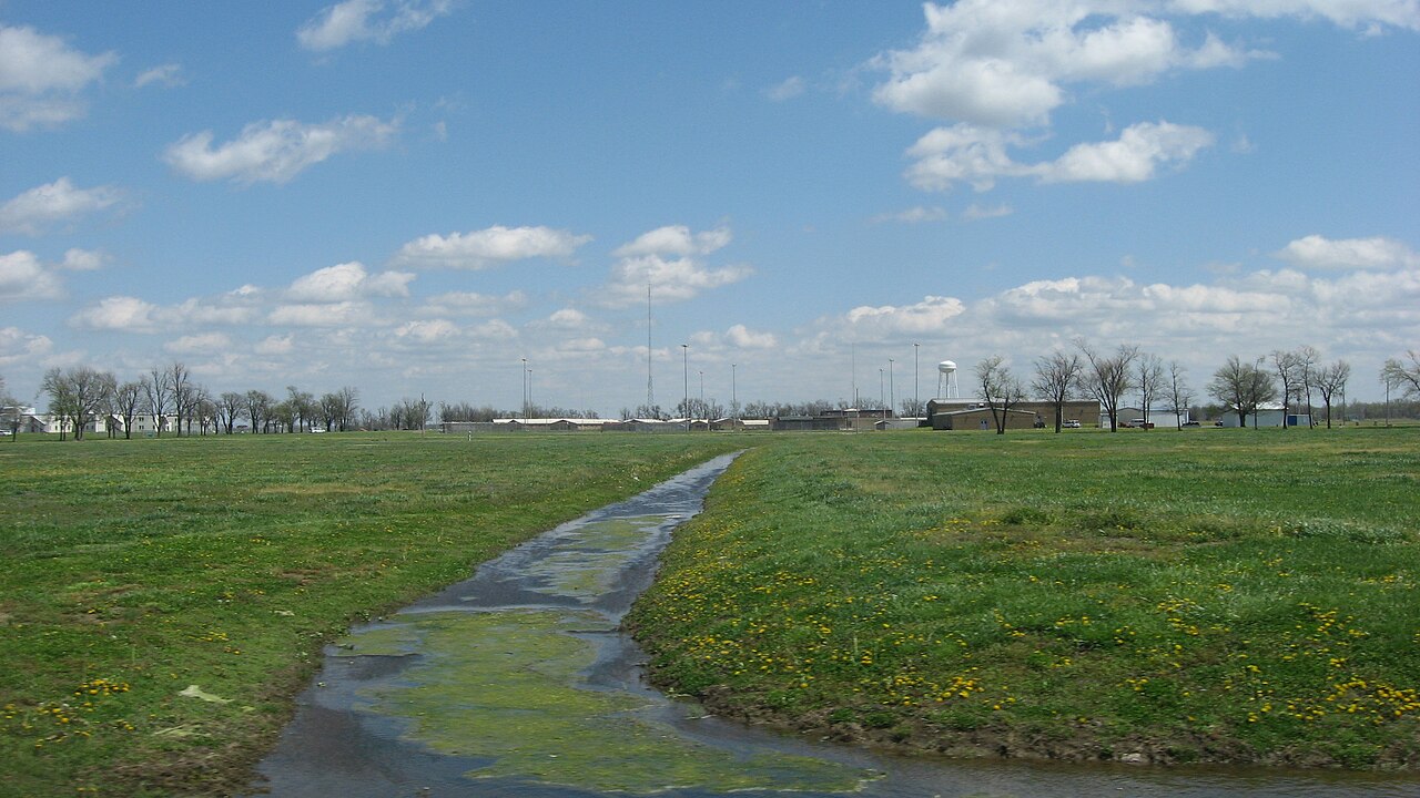 Northwest Correctional Complex seen across a grassy field and drainage ditch, with low buildings, towers and a water tower in the distance