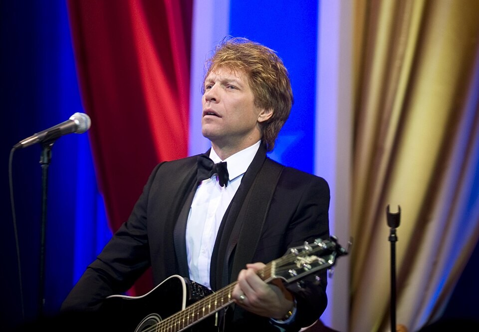 Jon Bon Jovi in a black tuxedo playing a black acoustic guitar beside a microphone, with colorful drapes behind him