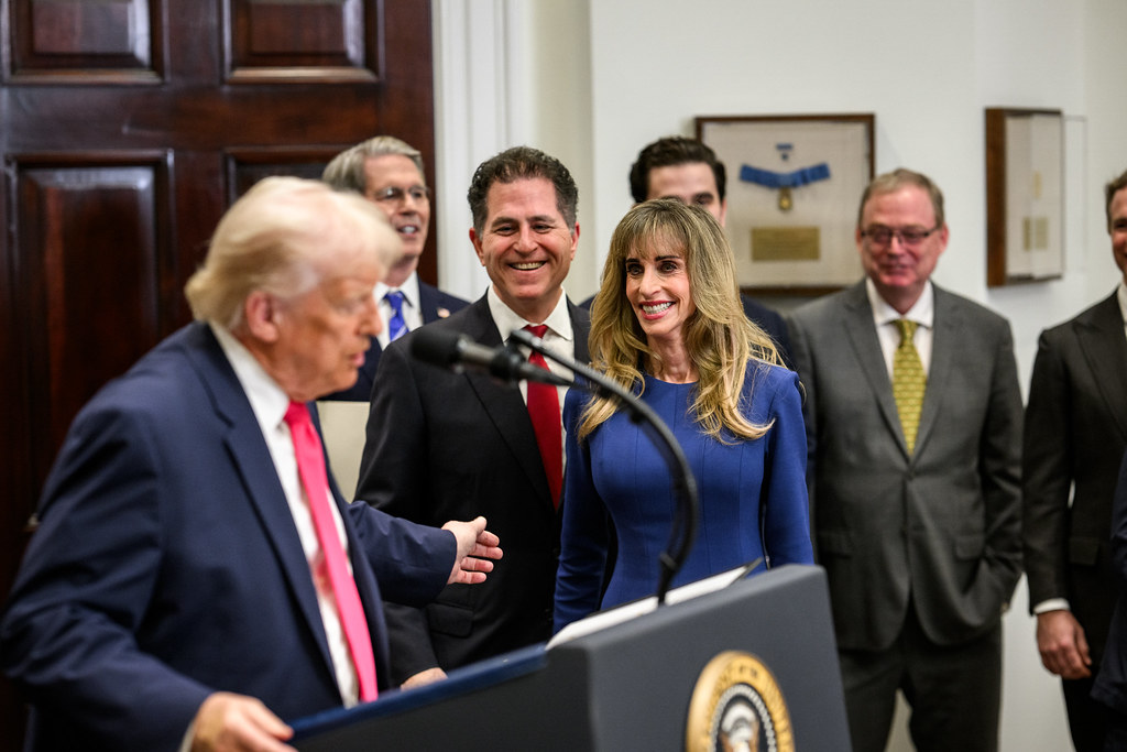 Susan Dell in a blue dress beside Michael Dell while Donald Trump gestures from a White House lectern