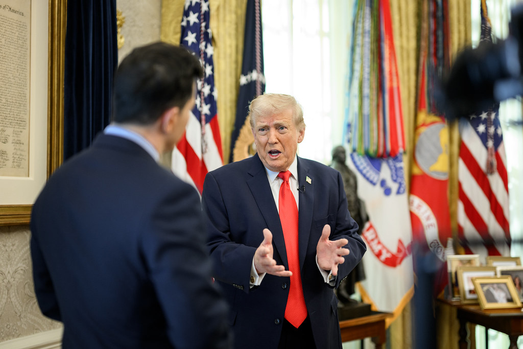 Donald Trump gestures while speaking with NBC anchor Tom Llamas in the Oval Office, with flags behind him