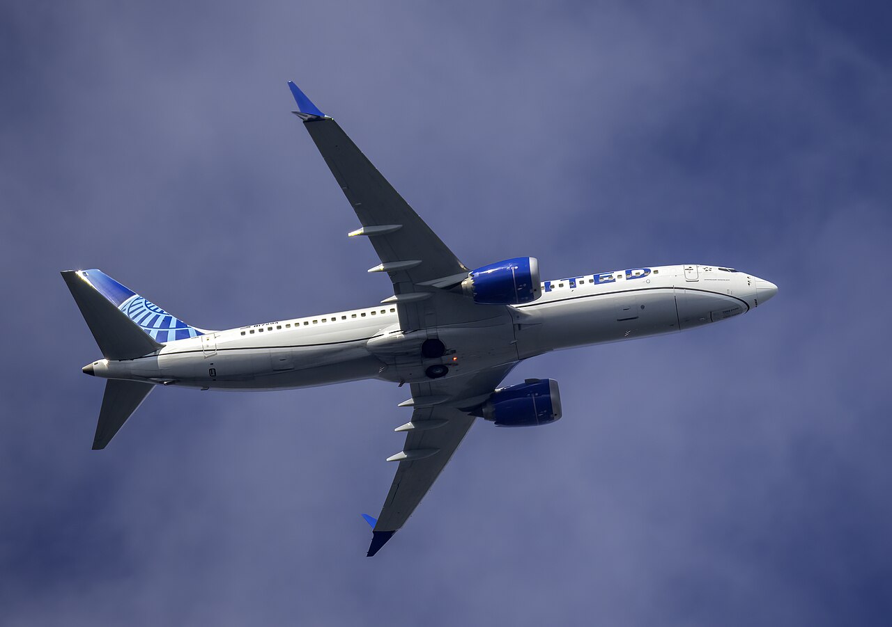 A United Airlines Boeing 737 MAX 8 photographed from below against a blue sky, with its landing gear extended